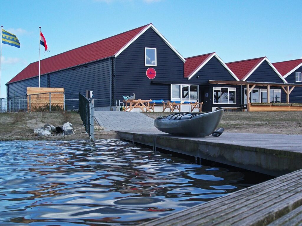 Het clubhuis van Kanovereniging Dolfijnen aan het water: donkerblauwe loodsen met rode daken, een steiger en een kano.
